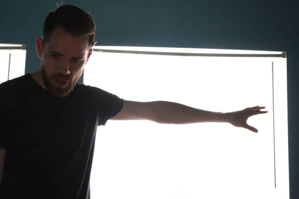 Screenshot of a scene from Mad Max. Features a guy wearing a metal hockey mask, speaking into a loudspeaker, with an army of post-apocalyptic soldiers and their vehicles on the desert slope behind him.