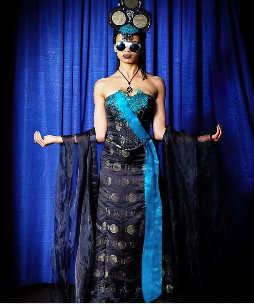 Queen of Clubs cosplay outfit by Chad Evett features a model dressed in black and turquoise with a headpiece sporting the infamous club symbol.