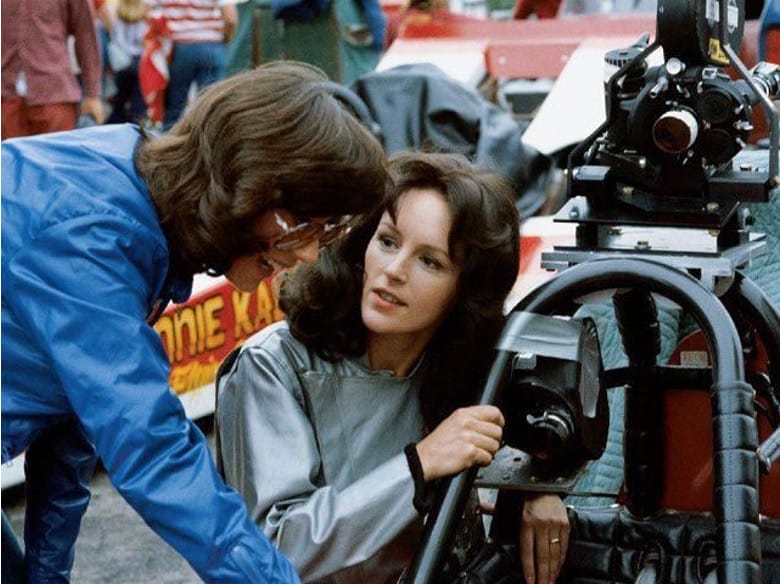 Image from Heart Like a Wheel, a 1983 movie, starring Beau Bridges, and written by Ken Friedman. Images shows 2 women standing near a movie camera, with 1980's hairdos and clothing that was symbolic of the era.