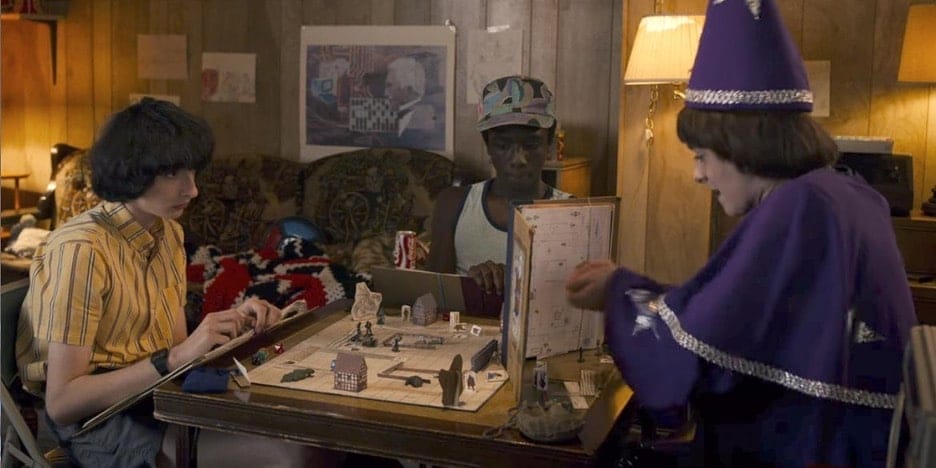 Image of 3 characters from the Netflix series: Stranger Things, playing a game of Dungeons and Dragons. The Dungeon Master is dressed in a purple wizard outfit.