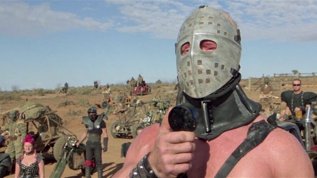 Screenshot of a scene from Mad Max. Features a guy wearing a metal hockey mask, speaking into a loudspeaker, with an army of post-apocalyptic soldiers and their vehicles on the desert slope behind him.