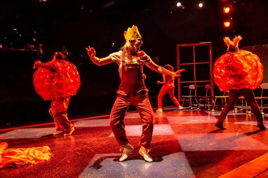 Still image of actors on stage, performing Lewis Carroll's Alice's Adventures in Wonderland for the theater. This is Bad Hats Theatre, on stage with chess board lighting, with a red hue.