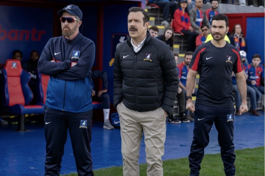 Image of the coaching staff from the Apple TV+ series: Ted Lasso. With characters left to right: Coach Beard, (Brendan Hunt), Ted Lasso (Jason Sudeikis) and Roy Kent (Brett Goldstein).