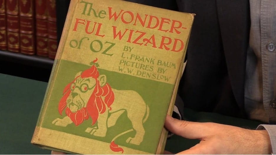 A person holding a book in a library. The old cover for "The Wonderful Wizard of Oz" by L. Frank Baum, with pictures by W.W. Denslow. Featuring a Lion, wearing glasses. 
