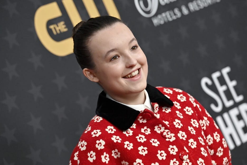 Image of Bella Ramsey, from The Last of Us and Game of Thrones fame. Here, she is smiling at the camera on the red carpet from a recent Hollywood premiere, wearing a red jacket with black collar and white floral pattern.