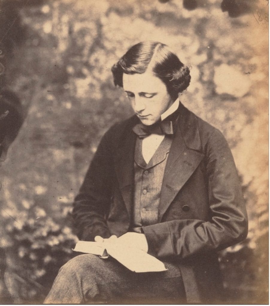 Old, black & white photograph of a young Lewis Carroll, wearing a suit and reading a book in the forest.