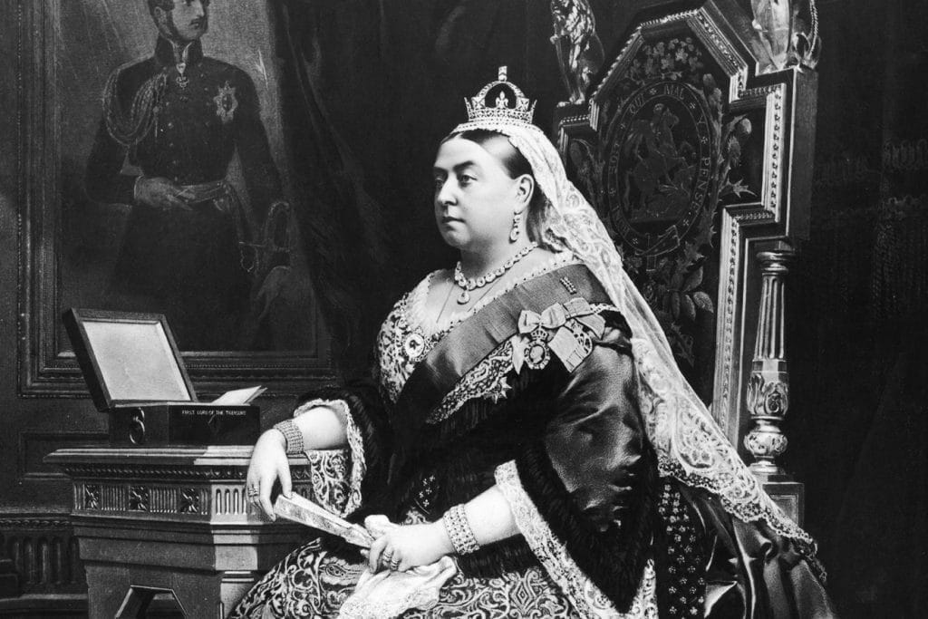 Old black & white photograph of England's Queen Victoria, wearing a crown and tiara, sitting on her throne. She was a massive inspiration to Lewis Carroll's Alice's Adventures in Wonderland.