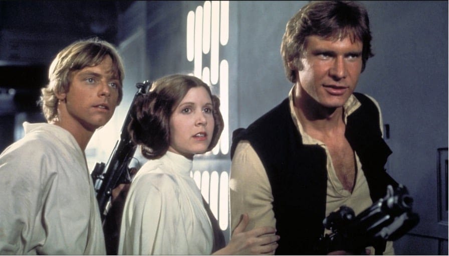 Image from the 1977 George Lucas film: Star Wars Episode IV: A New Hope. Pictured from left to right: Mark Hammil (Luke Skywalker), Carrie Fisher (Princess Leia Organa), Harrison Ford (Han Solo). They are all holding blasters and standing in a hallway on the Death Star.