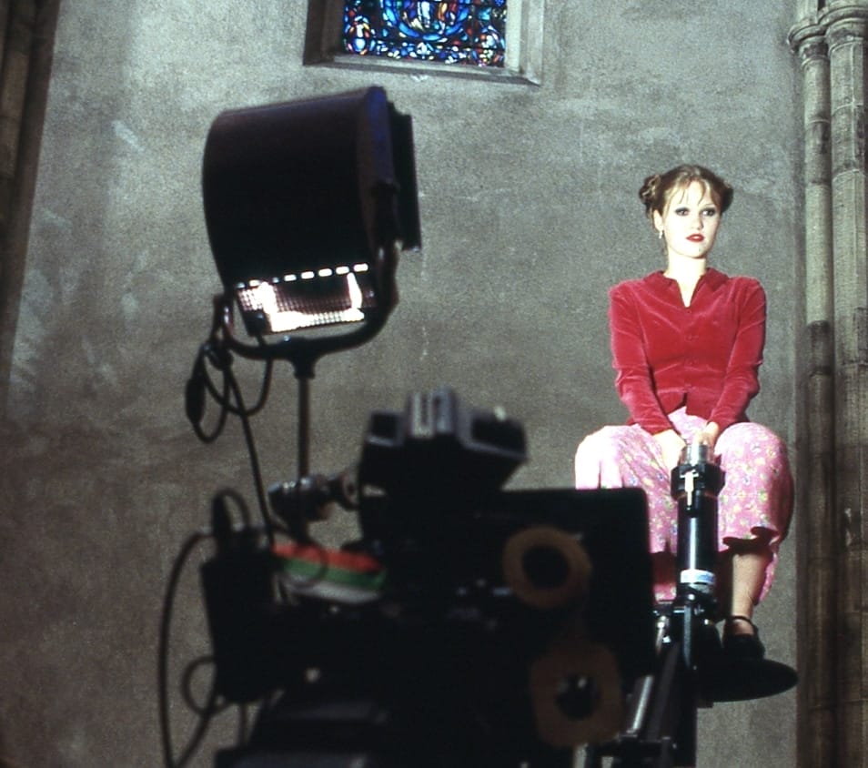 Behind-the-scenes image from the thriller movie "Wicked" featuring Julia Stiles, wearing a red top and pink skirt, sitting on a raised platform with a camera and light in the foreground.