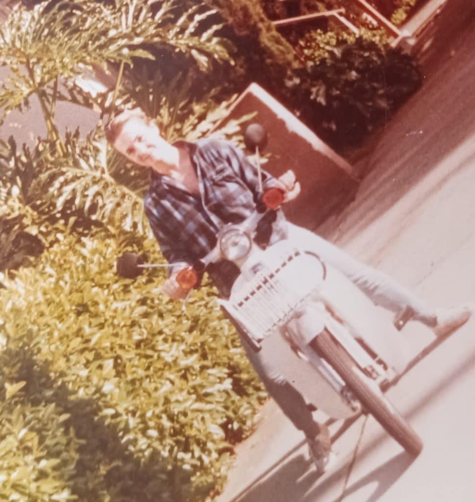 Author Liz Cavalier sitting on a white moped on a tree-lined sidewalk.