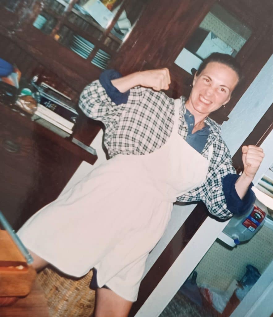 Author Liz Cavalier wearing a white apron and checkered shirt, posing with her fists raised.