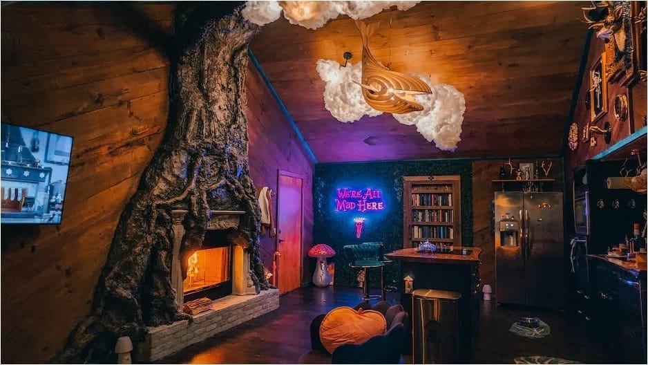 A living room and kitchen with wood-paneled walls and ceiling, a tree trunk-themed fireplace, and a neon sign reading "We're All Mad Here" from the Mad Hatter Cottage Airbnb in Mineral Bluff, Georgia.
