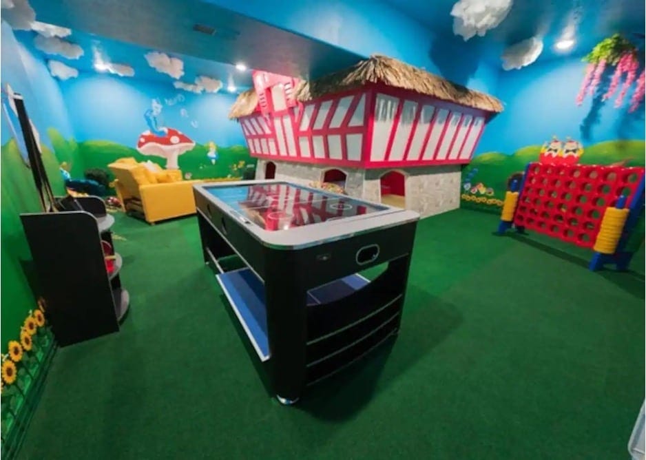 A playroom featuring an air hockey table, a large pink and white playhouse, and a blue and green landscape mural painted on the wall from the Alice in Wonderland Airbnb in Ozark, Missouri.