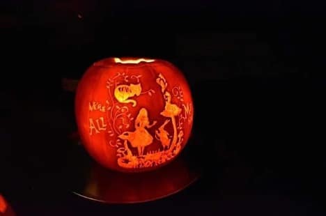 A photograph of a pumpkin carved to depict a scene from "Alice in Wonderland" with the quote "We're all mad here." 